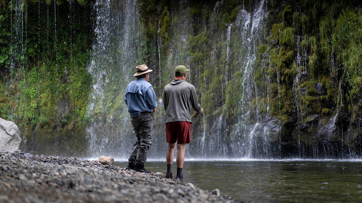 Two people standing by a waterfall, one holding a fishing rod.
